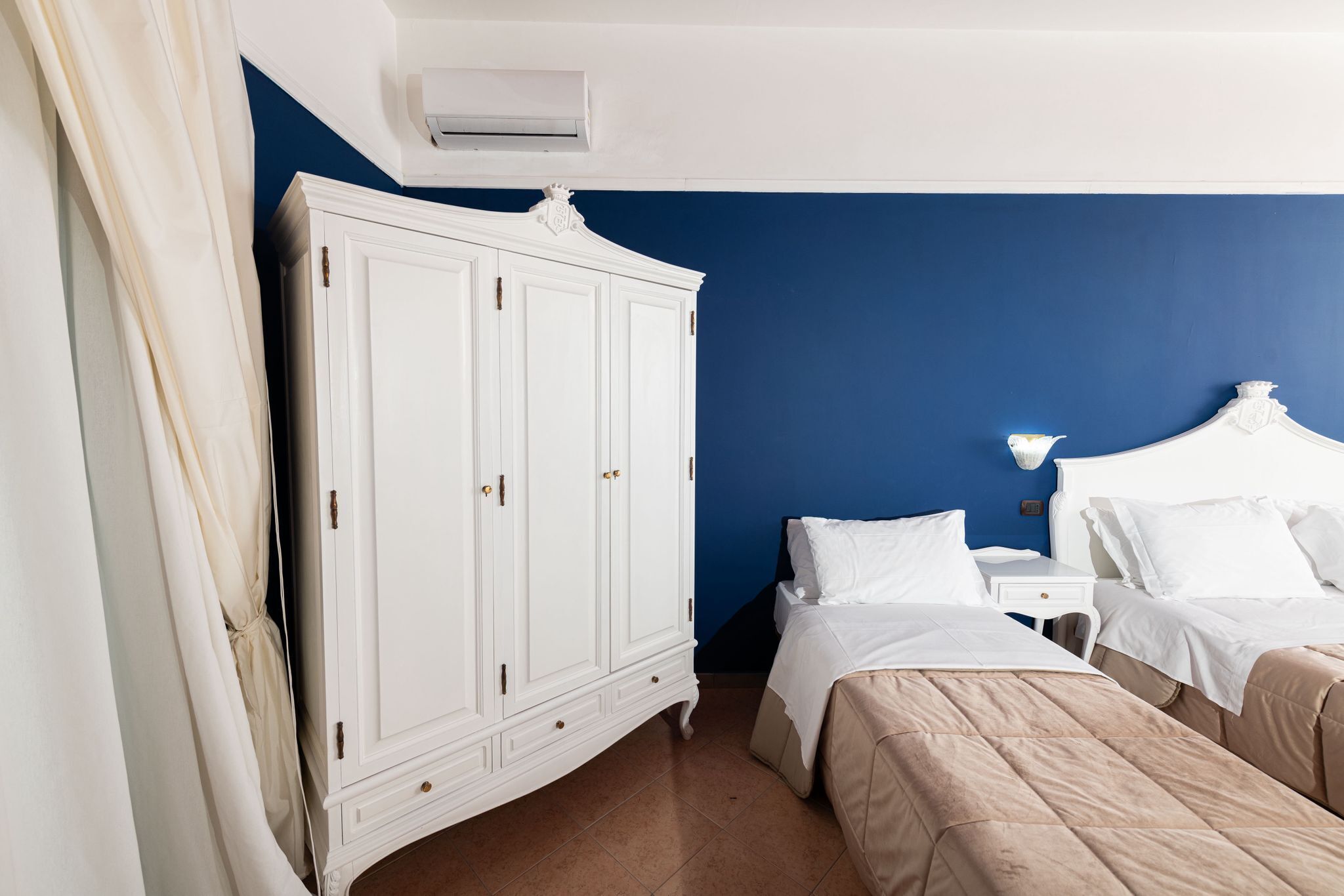 Un dormitorio con un armario blanco, dos camas, una mesita de noche y un aire acondicionado de pared.