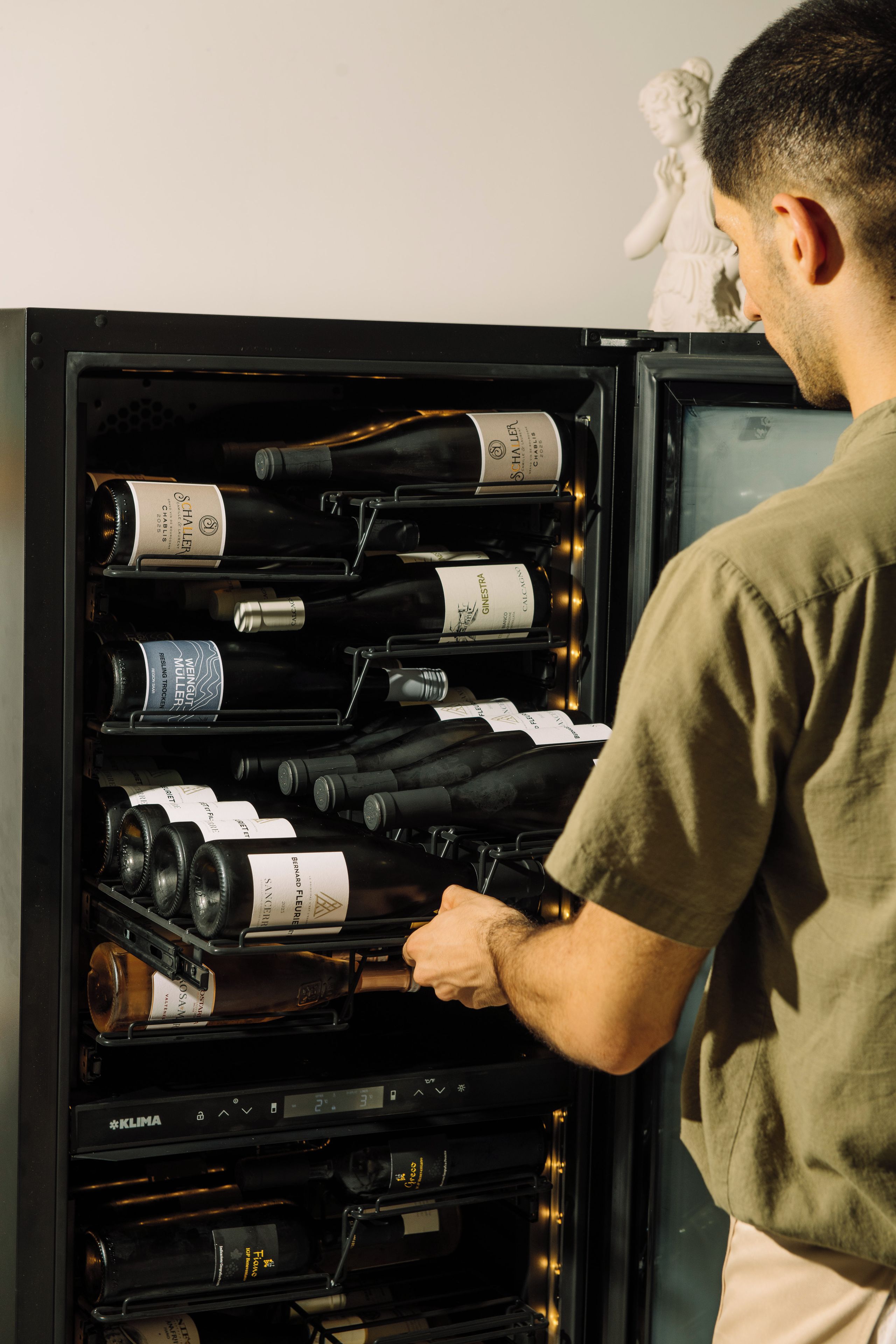 Un refrigerador de vinos repleto de botellas variadas, una persona alcanzando una botella y una estatua al fondo.