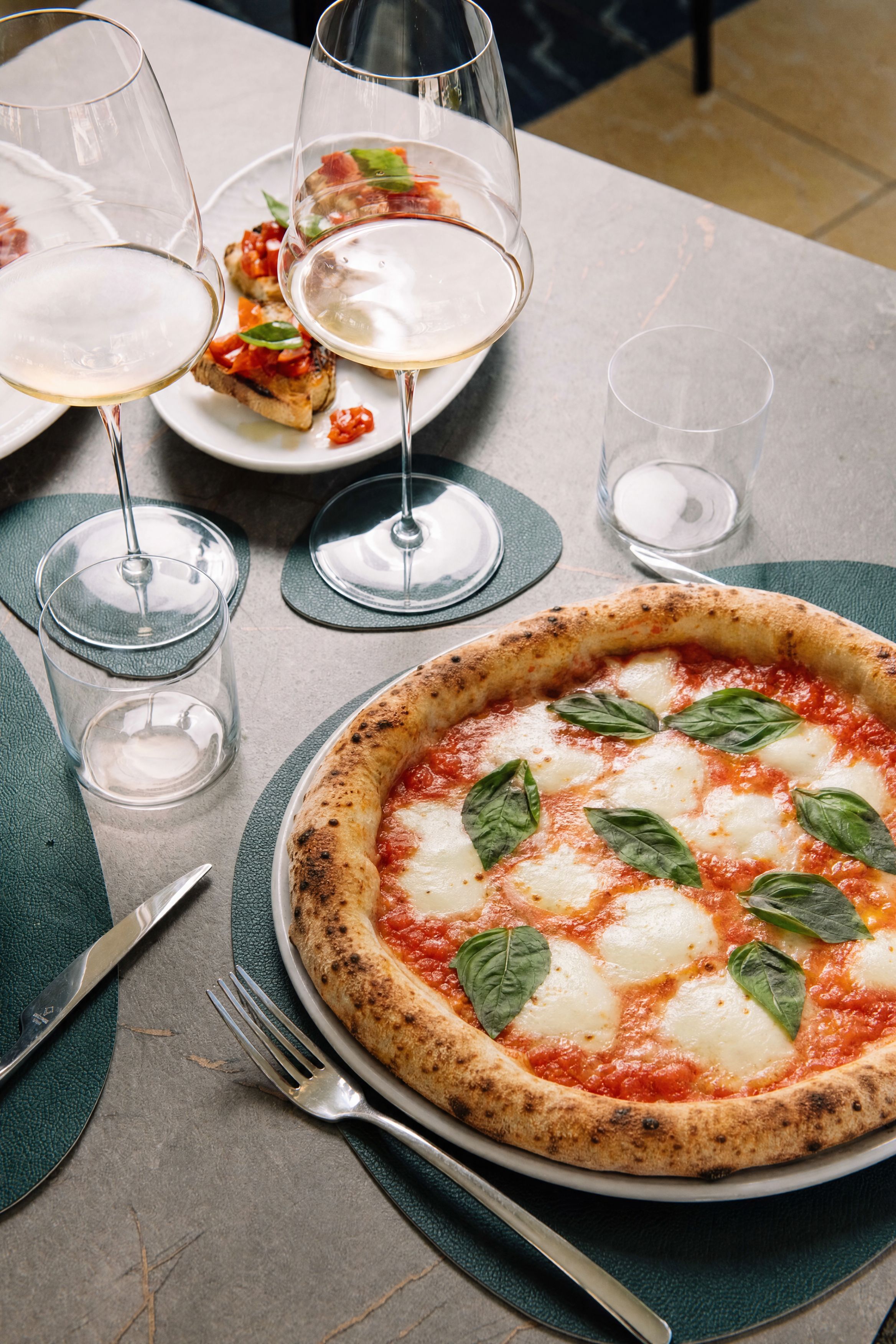 A pizza with basil leaves on a plate, accompanied by wine glasses and cutlery on a table.