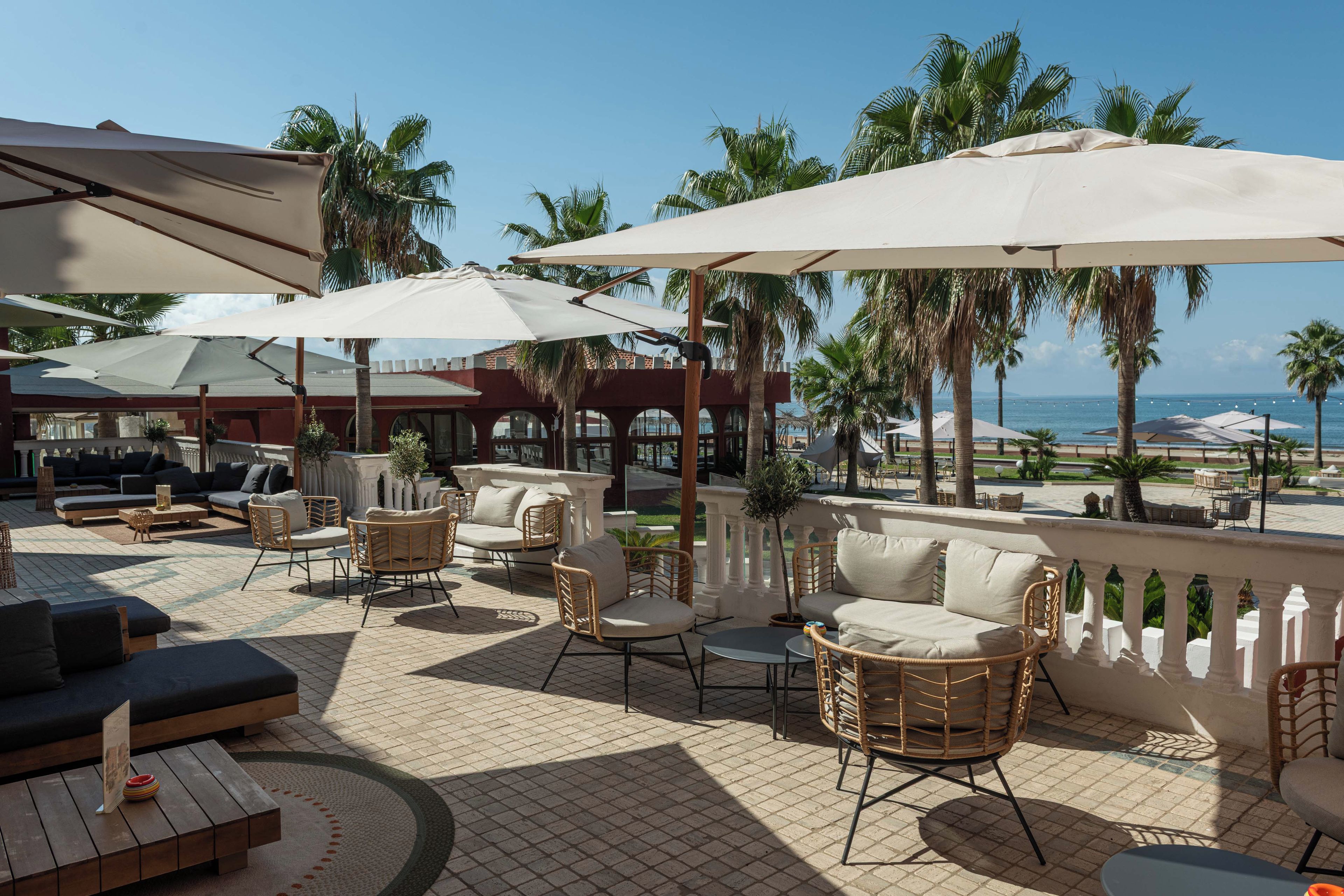 An outdoor seating area with wicker furniture and umbrellas, overlooking a beach with palm trees and a clear sky.