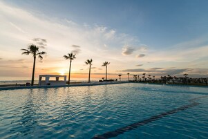 Una piscina con vistas al atardecer, palmeras y una playa al fondo.