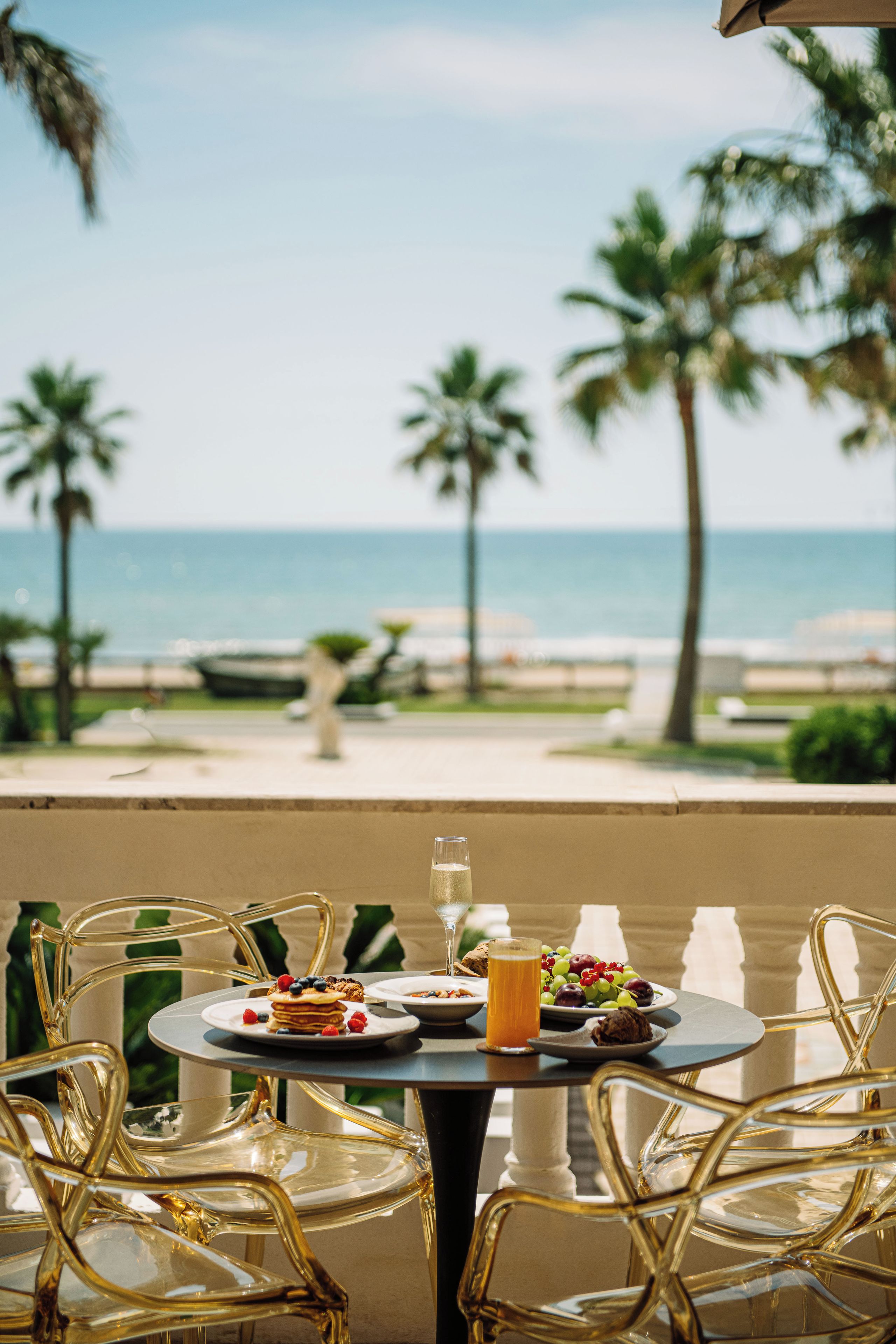 A table with breakfast items and a glass of orange juice, overlooking a beach with palm trees and the ocean.