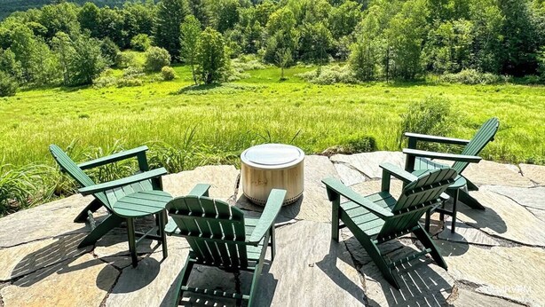 Un espacio de estar al aire libre con sillas Adirondack y una mesa redonda en un patio de piedra con vistas a un paisaje exuberante de árboles y colinas.