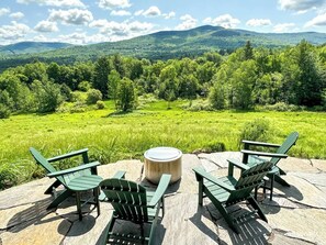 Ein Sitzbereich im Freien mit Adirondack-Stühlen und einem runden Tisch auf einer Steinterrasse mit Blick auf eine üppige grüne Landschaft aus Bäumen und Hügeln.