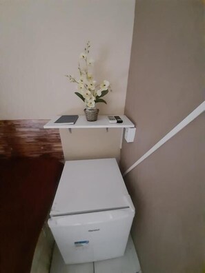 Una habitación pequeña con un refrigerador blanco, una estantería blanca con un jarrón de flores y un control remoto.