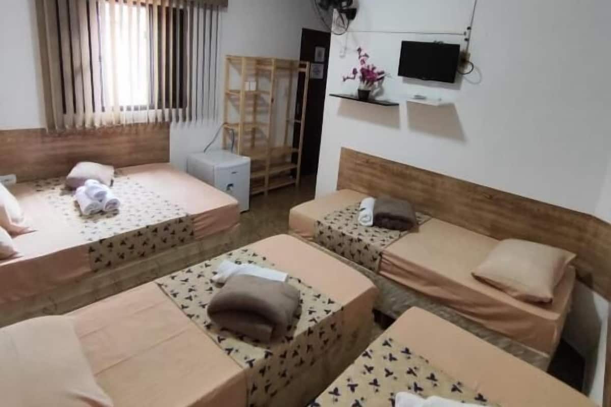 A room with four single beds, a wooden headboard, a TV mounted on the wall, and a window with blinds.