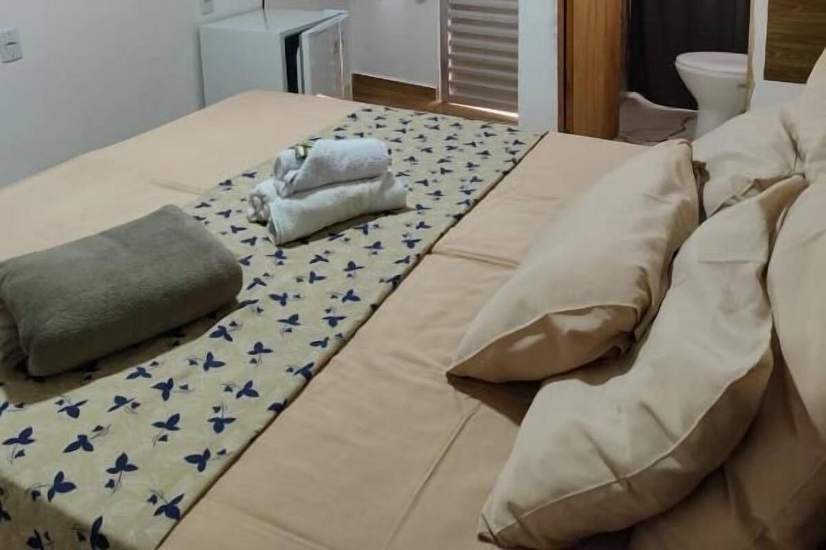 A bedroom with a bed, a bedside table, an air conditioning unit, a window with curtains, and a door leading to another room.