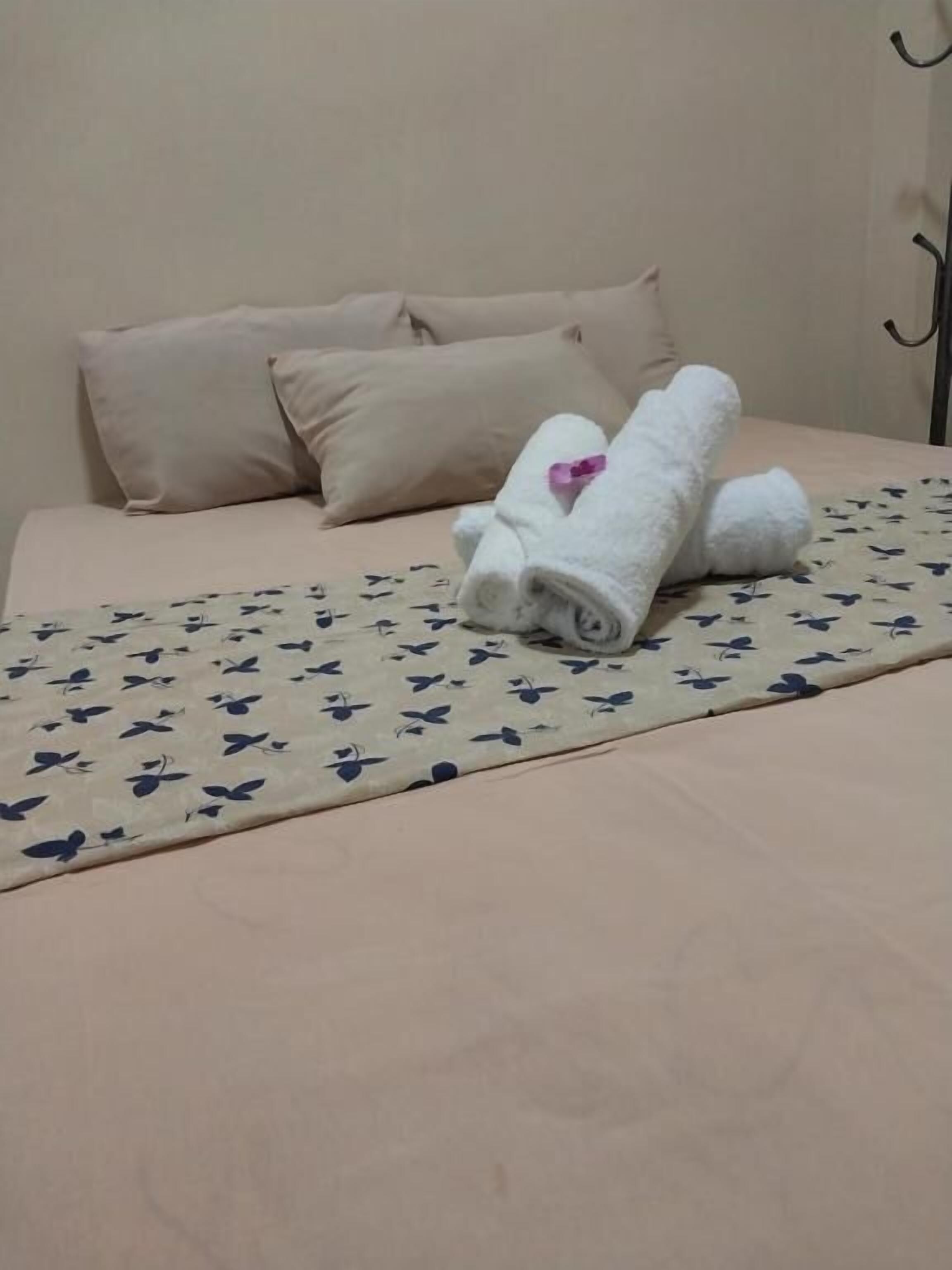 Un lit recouvert d’une couvre-lit à motifs floraux, deux oreillers beige et une serviette blanche roulée ornée d’une fleur rose.