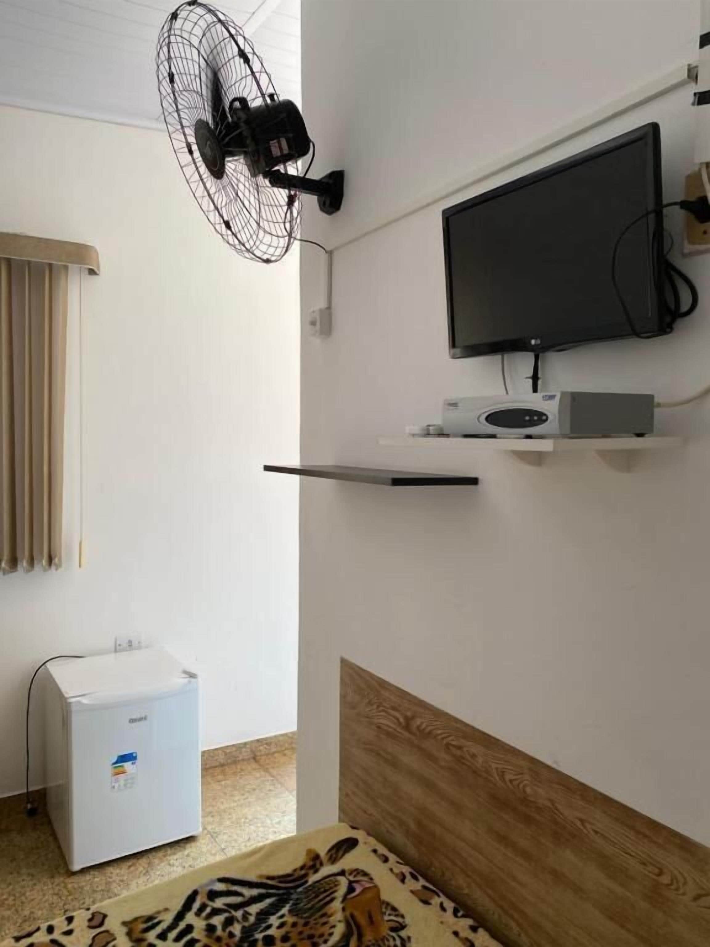 A room with a mounted TV, a ceiling fan, a small refrigerator, and a wooden headboard.