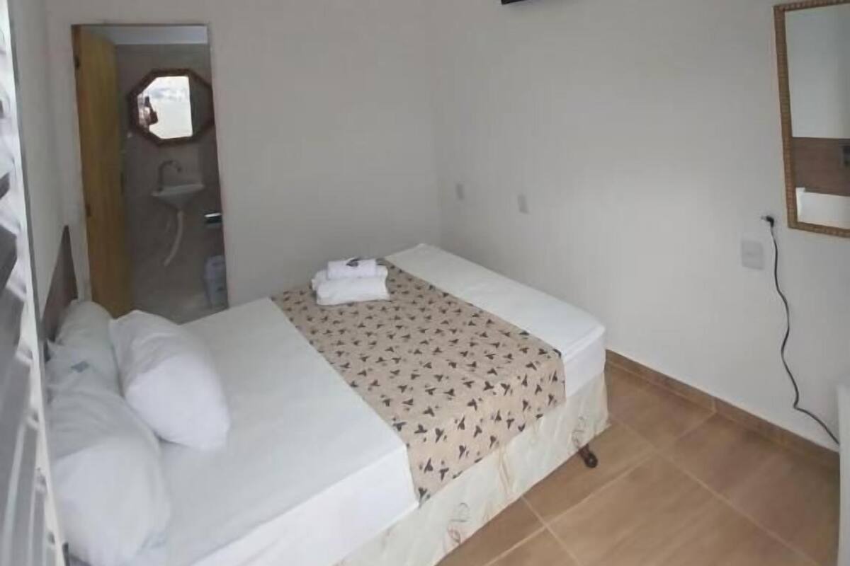A single bed with a white mattress, a patterned bedspread, a ceiling fan, a wall-mounted TV, and a tiled floor.