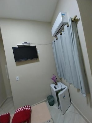 A small room with a TV mounted on the wall, a mini-fridge, a small table with a plant, and a red chair.
