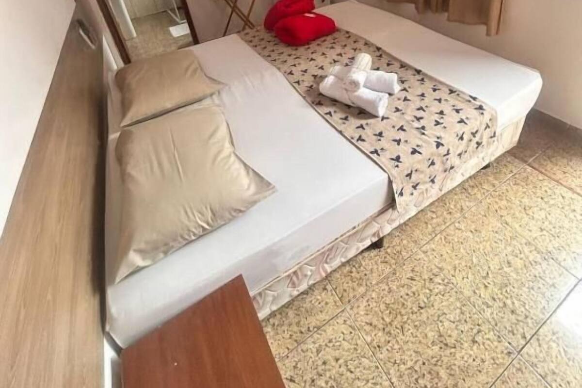 A single bed with a white mattress and beige pillows, a wooden nightstand, a red chair, a standing fan, and a window with beige curtains.