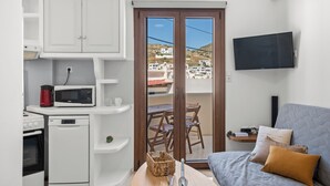 A compact living space with a kitchenette, a flat-screen TV, a sofa with cushions, and a view of buildings through a glass door. - Ageri House | Family Home in Vivlos, Naxos | Sleeps 6 (Vivlos)