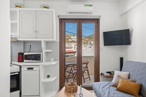 A compact living space with a kitchenette, a flat-screen TV, a sofa with cushions, and a view of buildings through a glass door. - Ageri House | Family Home in Vivlos, Naxos | Sleeps 6 (Vivlos)