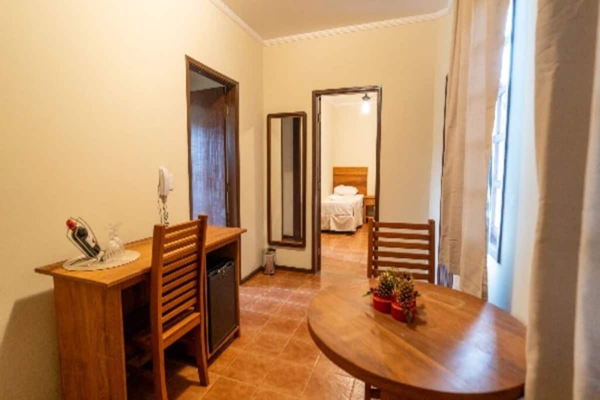 A hotel room with a wooden desk, chairs, a round table with a plant, a mirror, and a bed with a white bedspread.