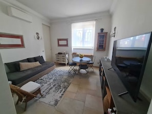 Ein kleiner Wohnbereich mit einer Couch, einem Esstisch mit Stühlen, einem Fernseher und einem Fenster.