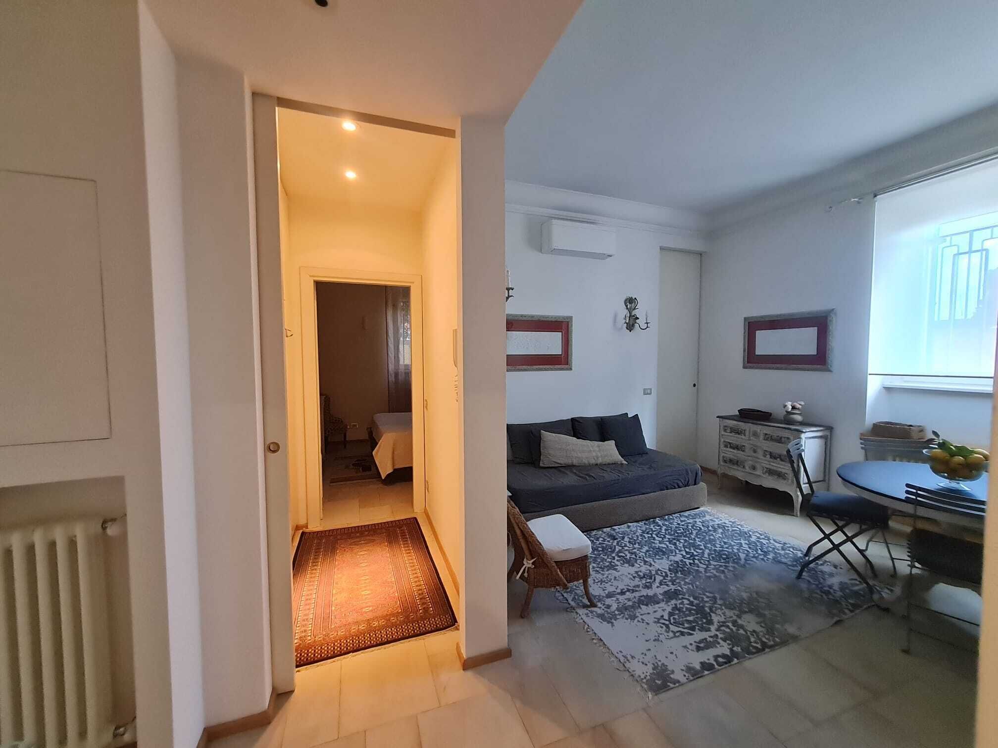 A compact living space with a sofa, a small dining table, a chair, a radiator, and a door leading to another room.