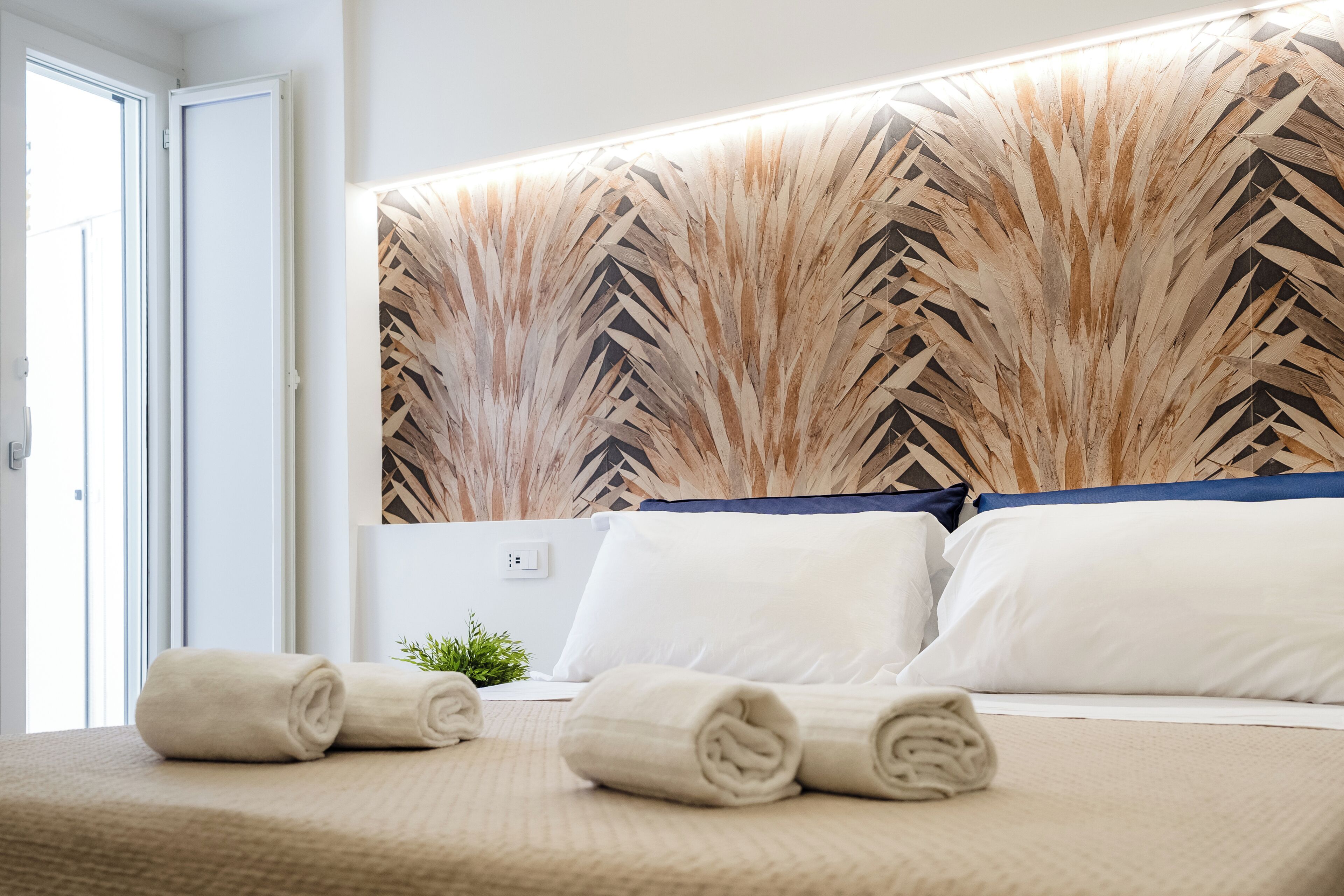 Une chambre d’hôtel avec un lit, deux serviettes enroulées, une petite plante et un mur orné d’un motif géométrique.