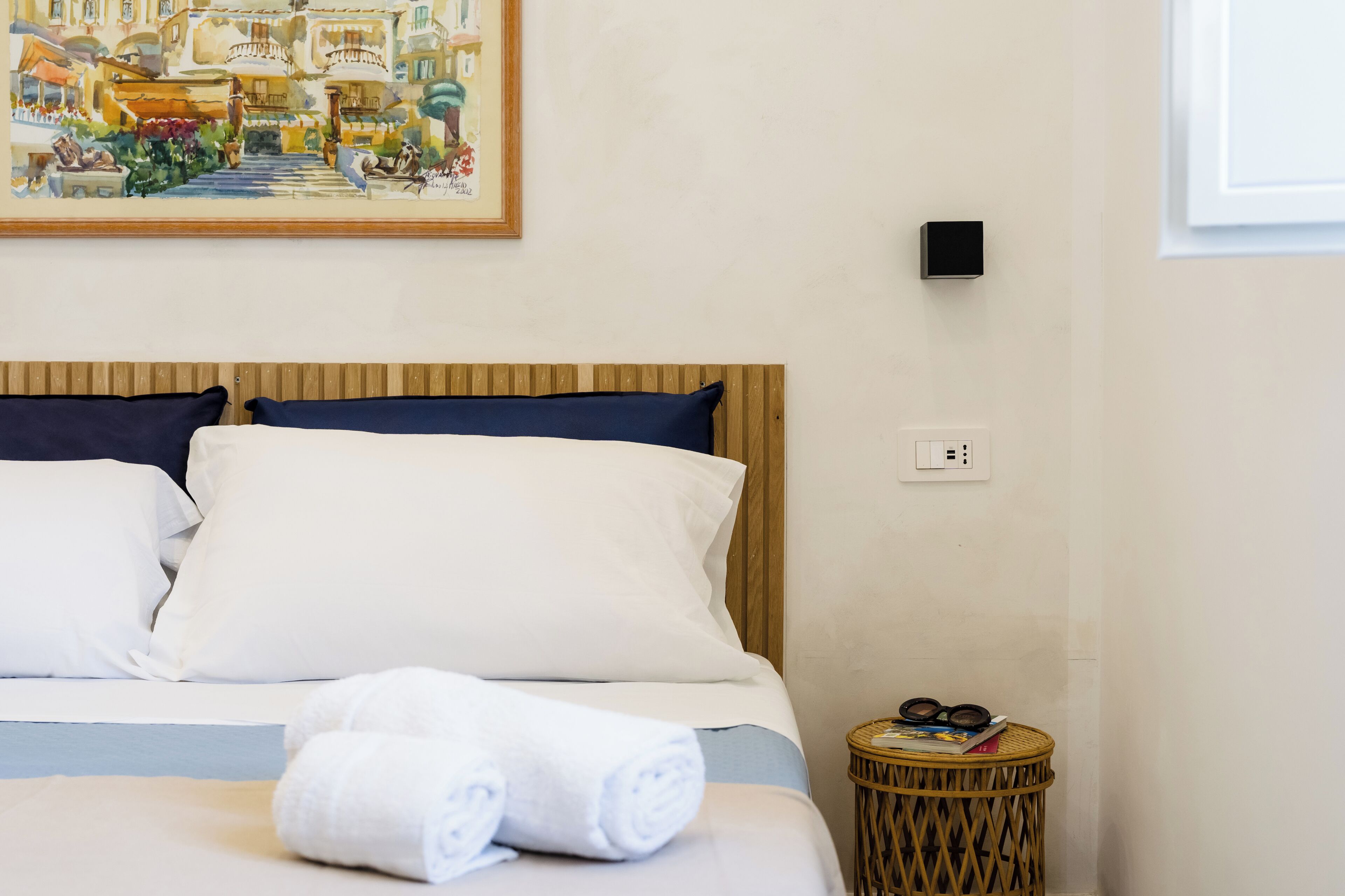 Une chambre d’hôtel avec un lit, des oreillers blancs et bleu marine, une serviette blanche enroulée, une petite table d’appoint ronde en osier avec un livre et des lunettes, et un tableau représentant une scène de rue accroché au mur.