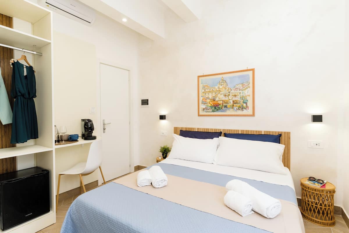 A hotel room with a bed, a chair, a wardrobe, a small table with a plant, and a painting on the wall.