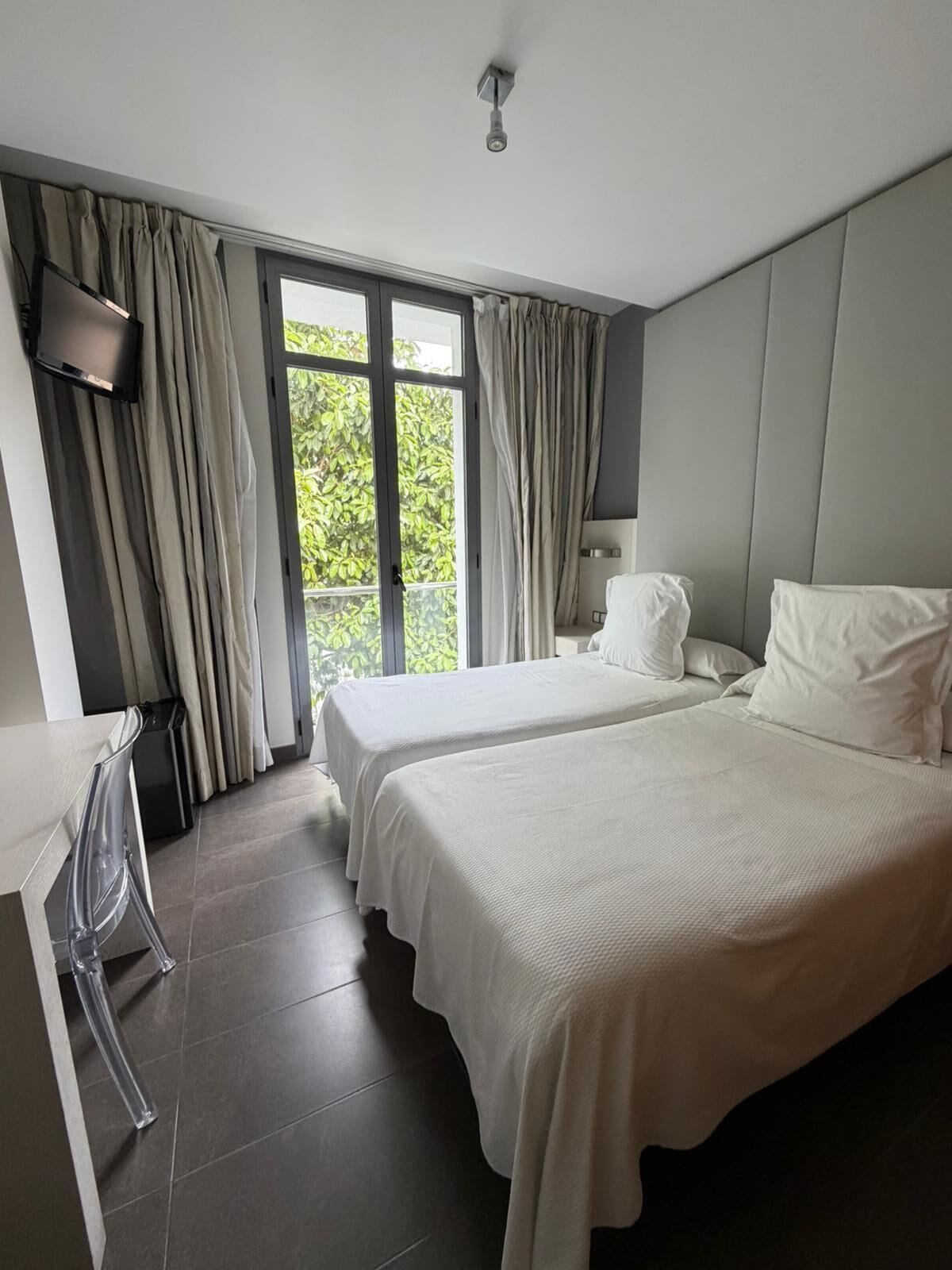 A hotel room with two beds, a desk, a TV, and a window with curtains.