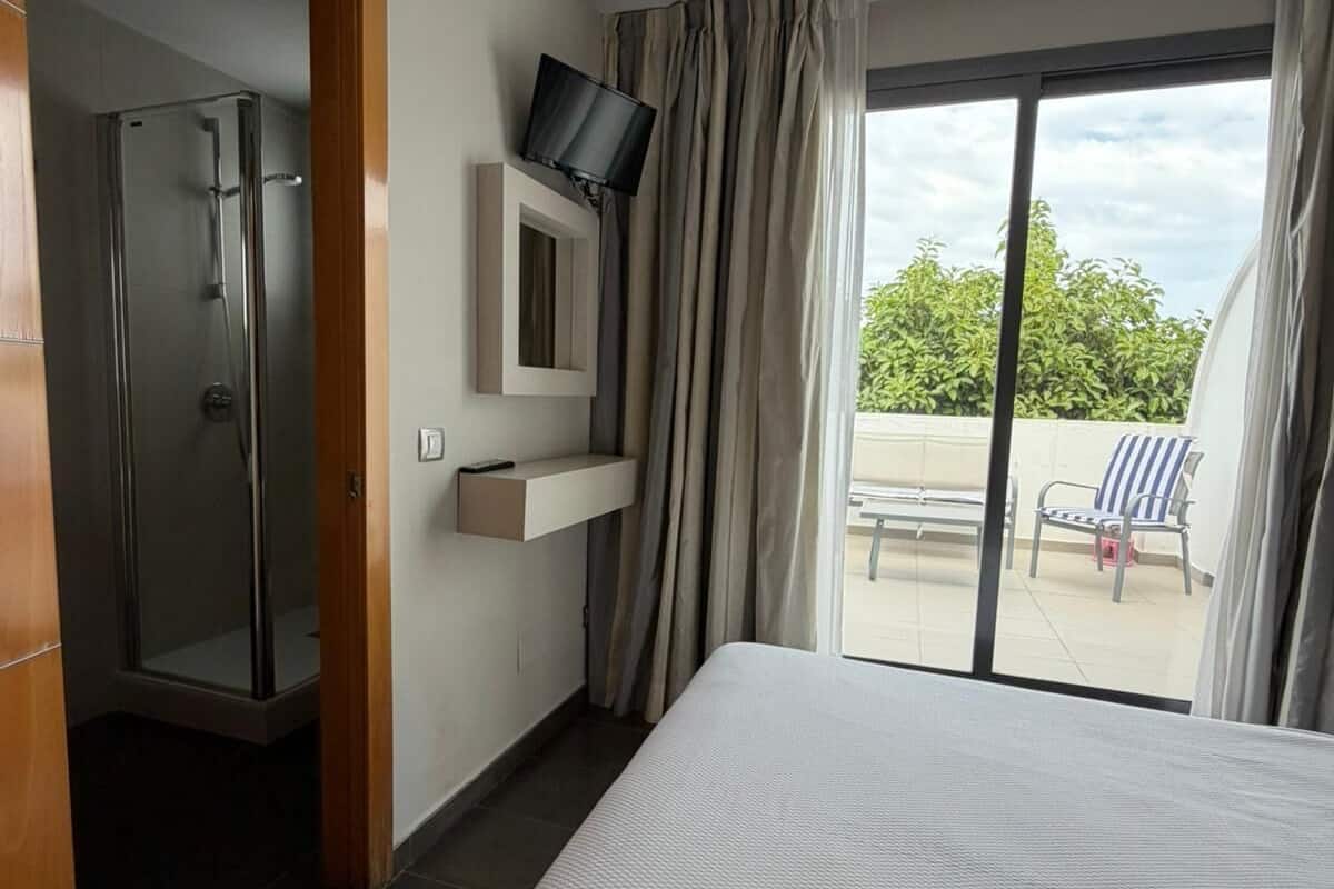 A hotel room with a bed, a shower, a TV mounted on the wall, and a balcony with chairs.