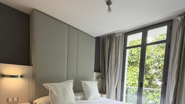 Une chambre à coucher moderne avec un grand lit, une table de chevet, une lampe fixée au mur et une fenêtre avec des rideaux.