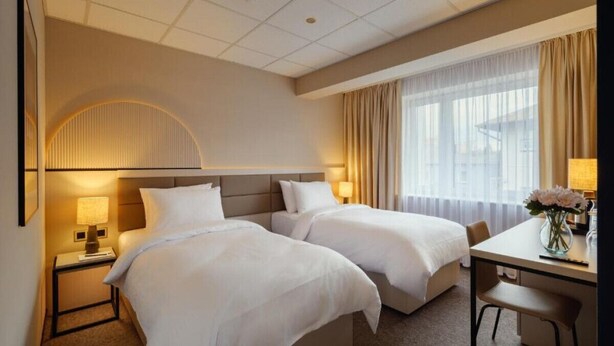 A hotel room with two beds, a desk, a chair, a lamp, and a window with curtains. - Yuko Residence (Jasło)