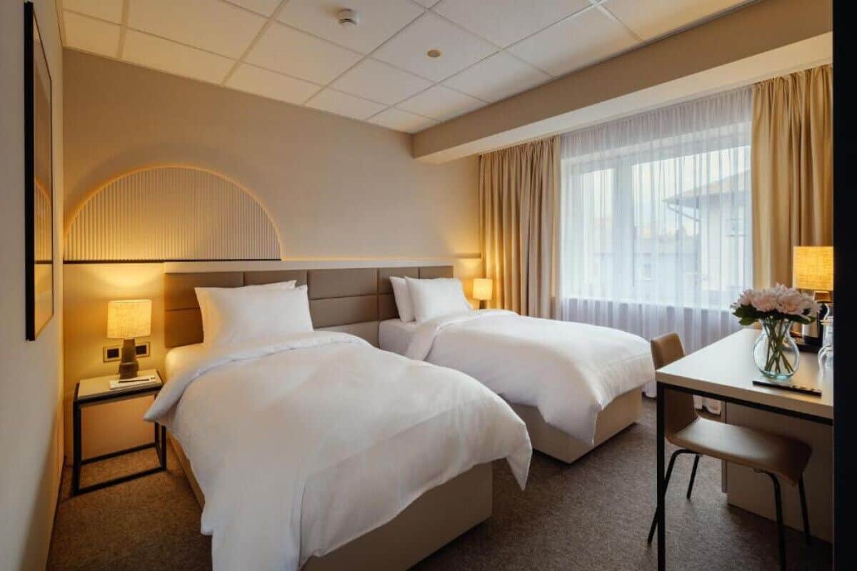 A hotel room with two beds, a desk, a chair, a lamp, and a window with curtains.