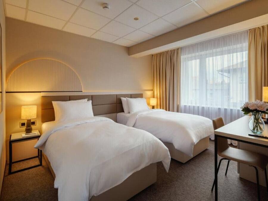Habitación de hotel con dos camas, un escritorio, una silla, una lámpara y una ventana con cortinas.