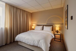 A hotel room with a large bed, two bedside tables with lamps, a headboard with a light fixture, and a large window with curtains.
