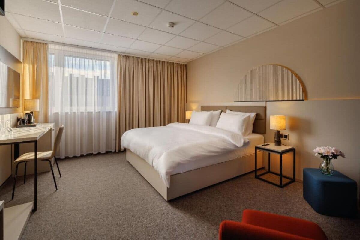 A hotel room with a bed, bedside tables, a desk, a chair, and a small table with a vase of flowers.