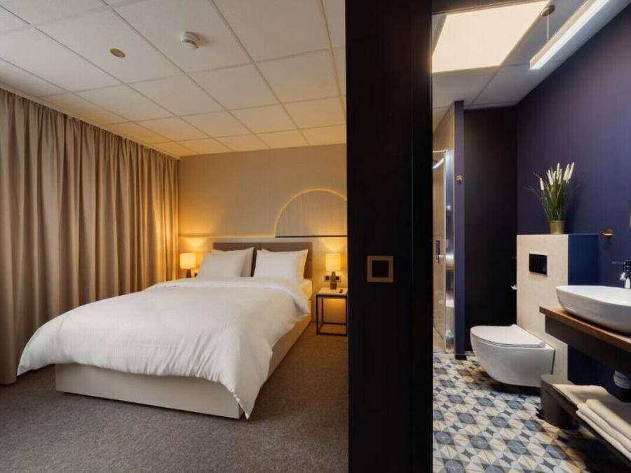 Habitació d'hotel amb un llit, una tauleta de nit, una làmpada i un bany amb un lavabo i un vàter.