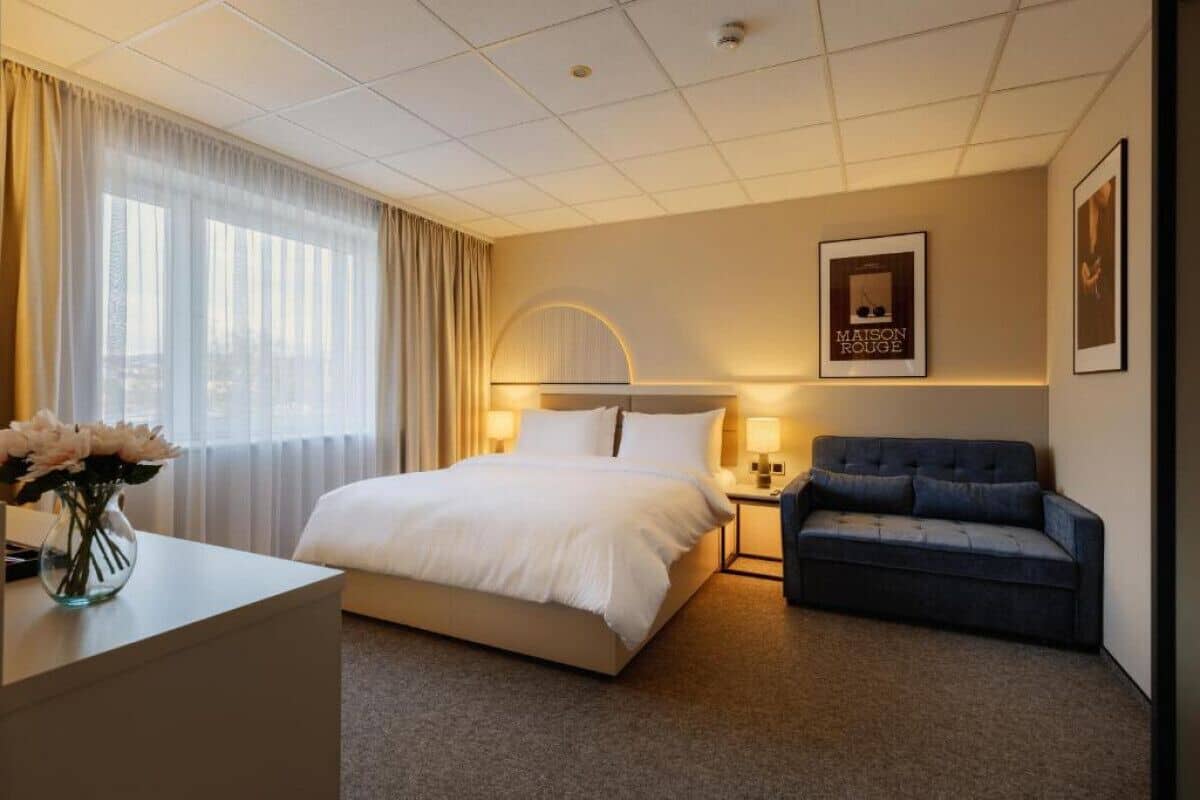 A hotel room with a bed, bedside table, lamp, and a sofa.