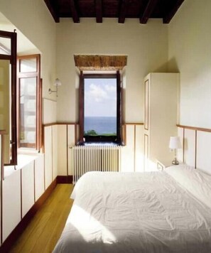 A bedroom with a large bed, a window with a view of the sea, a radiator, and wooden beams on the ceiling. - Apartment LEKEITIO terrace, garden and sea views (Bizkaia)
