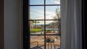 A view through a window with a metal grid, a patio area with a table and chairs, and a landscaped garden with a palm tree.