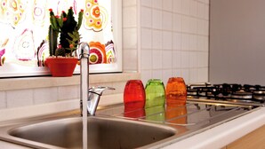 A kitchen sink with running water, a colorful window decoration, and a stove in the background. - CrauNari Agritourism and Educational Company (Modica)