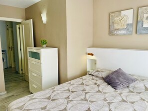 Een slaapkamer met een bed, een dressoir, een nachtkastje, een wandlamp en een deur naar een andere kamer.