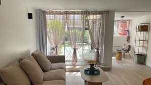A living room with a sofa, coffee table, and a view through sheer curtains to an outdoor area with a table and chairs. - Villa Mandarine (Arcachon)