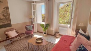 A room with a red sofa, a wicker chair, a small table, a potted plant, and a window with curtains. - La maison du repos (Hyères)