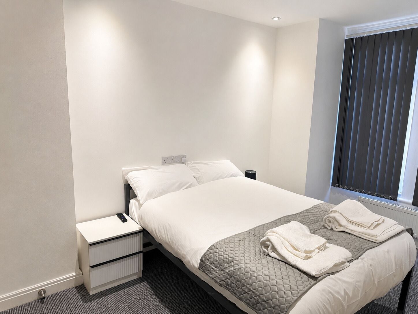 A hotel room with a bed, bedside table, and a window with blinds.