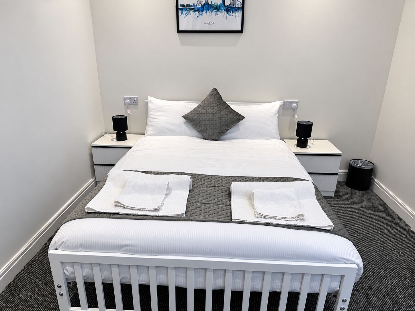 Une chambre d’hôtel avec un lit, deux tables de chevet, une poubelle et un tableau encadré au mur.