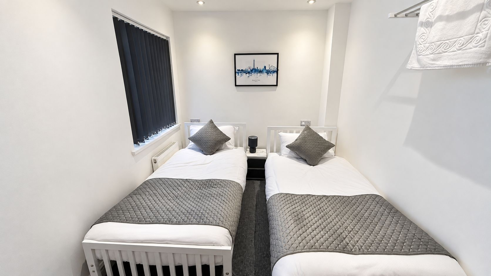 A twin room with two beds, a window with blinds, a nightstand with a lamp, and a framed picture on the wall.