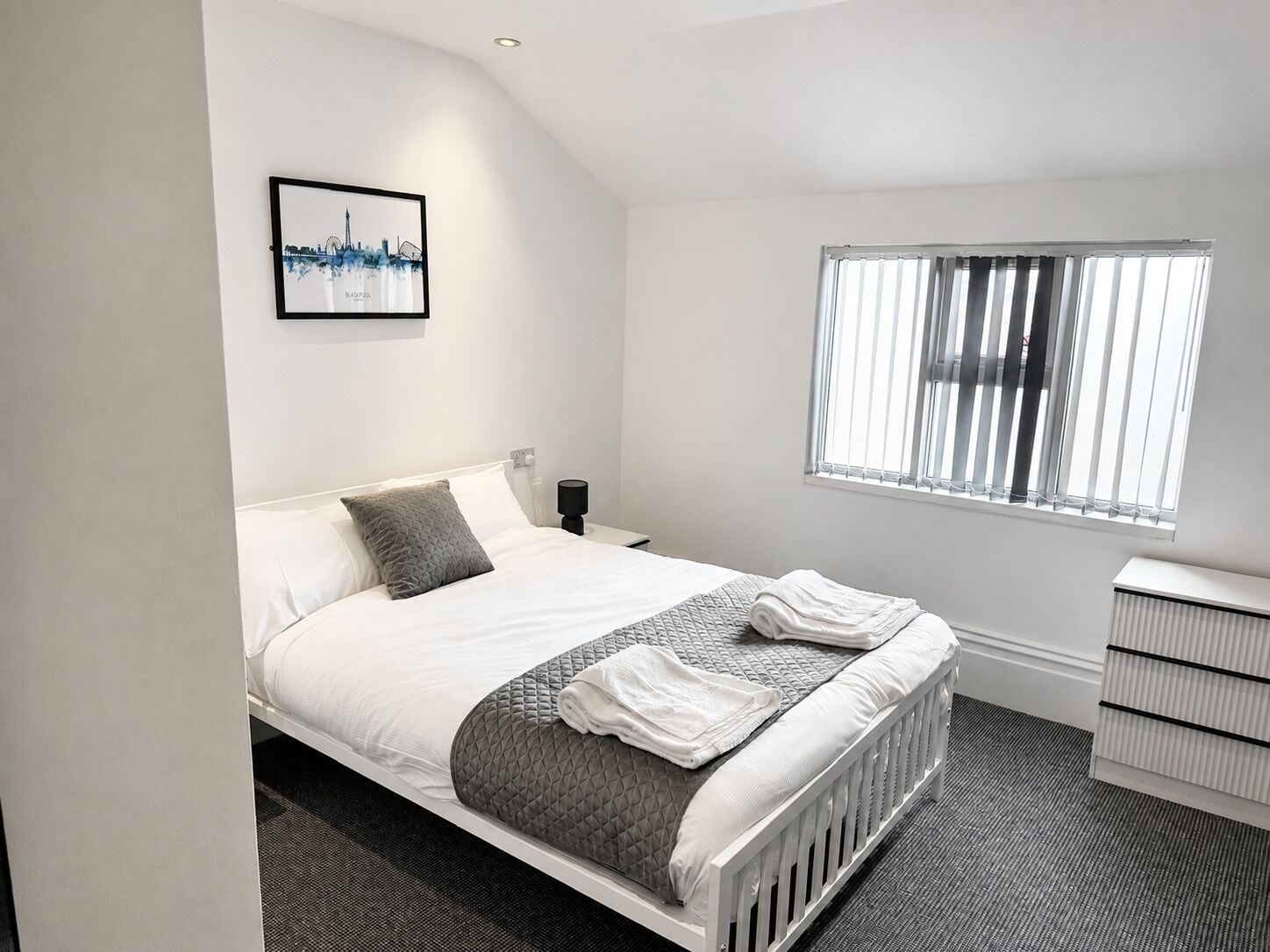 A bedroom with a bed, a bedside table, a nightstand, a window with blinds, and a framed picture on the wall.