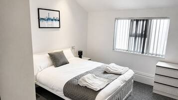 A bedroom with a bed, a bedside table, a nightstand, a window with blinds, and a framed picture on the wall.