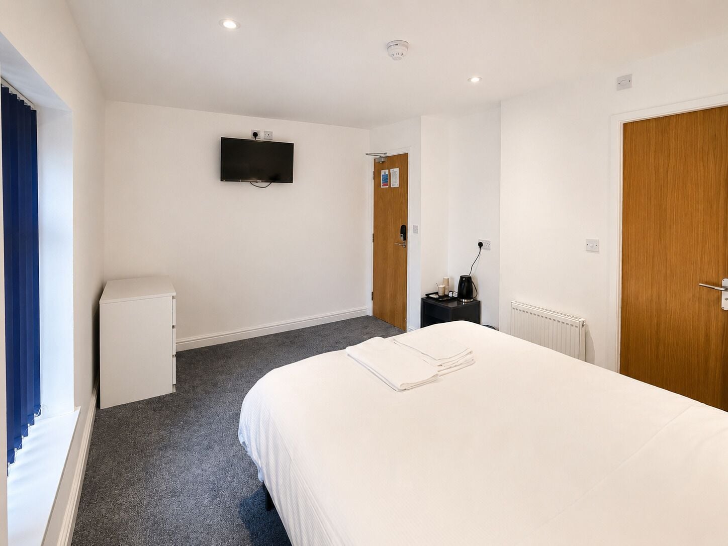 A hotel room with a bed, a television mounted on the wall, a small refrigerator, and a wooden door.