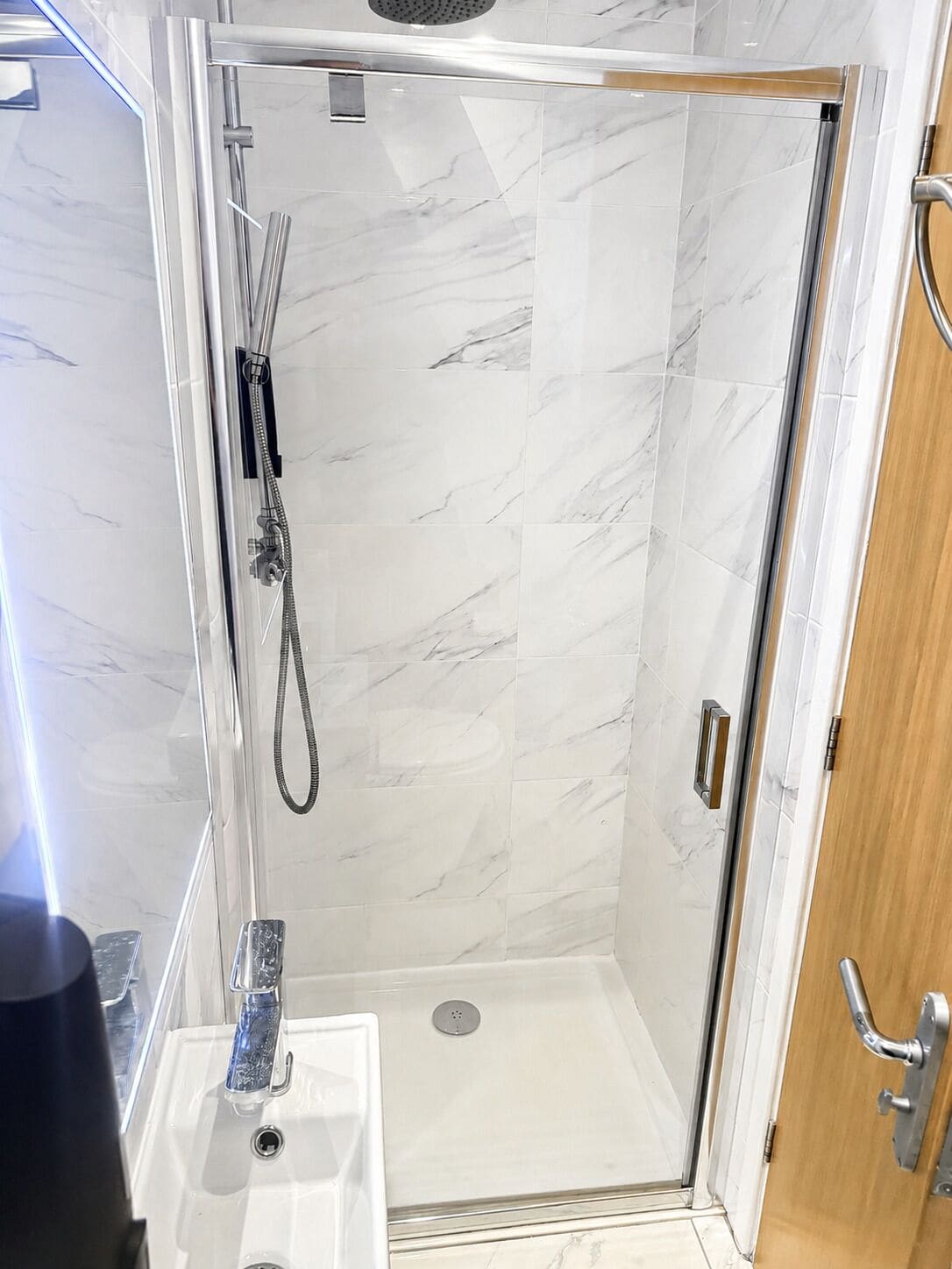 A glass-enclosed shower with a marble wall, a sink with a faucet, and a wooden door.