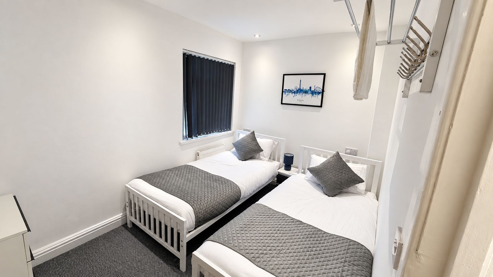 A hotel room with two beds, a window with blinds, a bedside table with a lamp, and a wall-mounted artwork.
