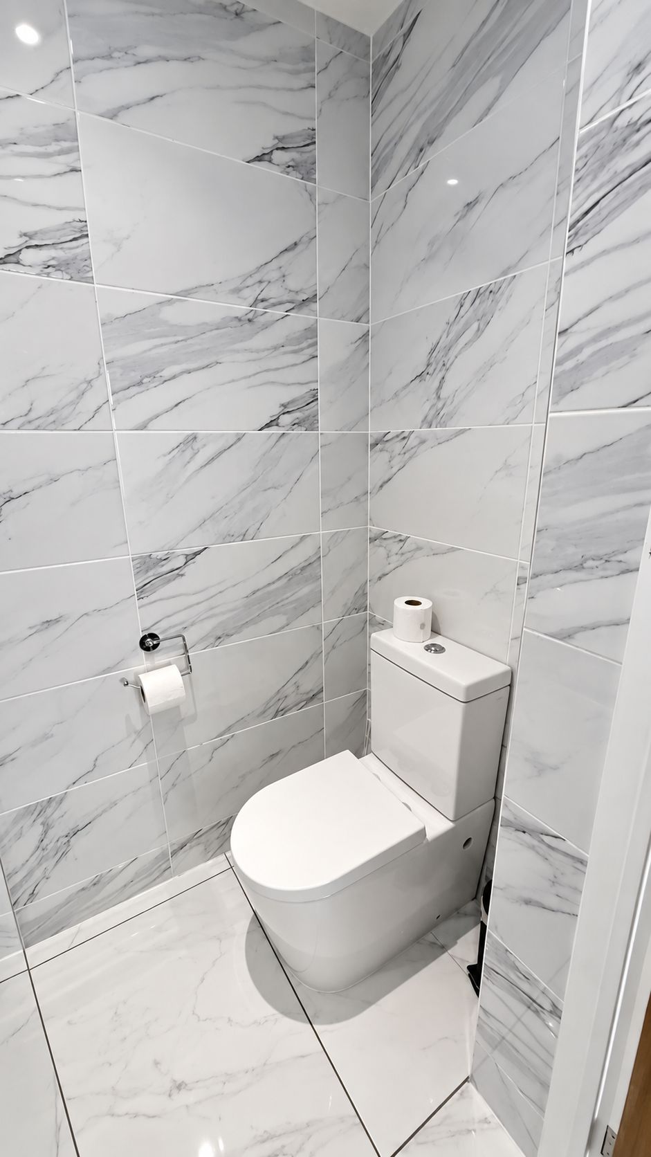 A bathroom with a toilet, marble tiles, and a roll of toilet paper on the wall.