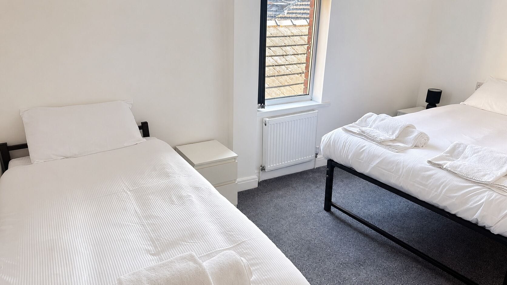A bedroom with two beds, a window, a radiator, and a nightstand.
